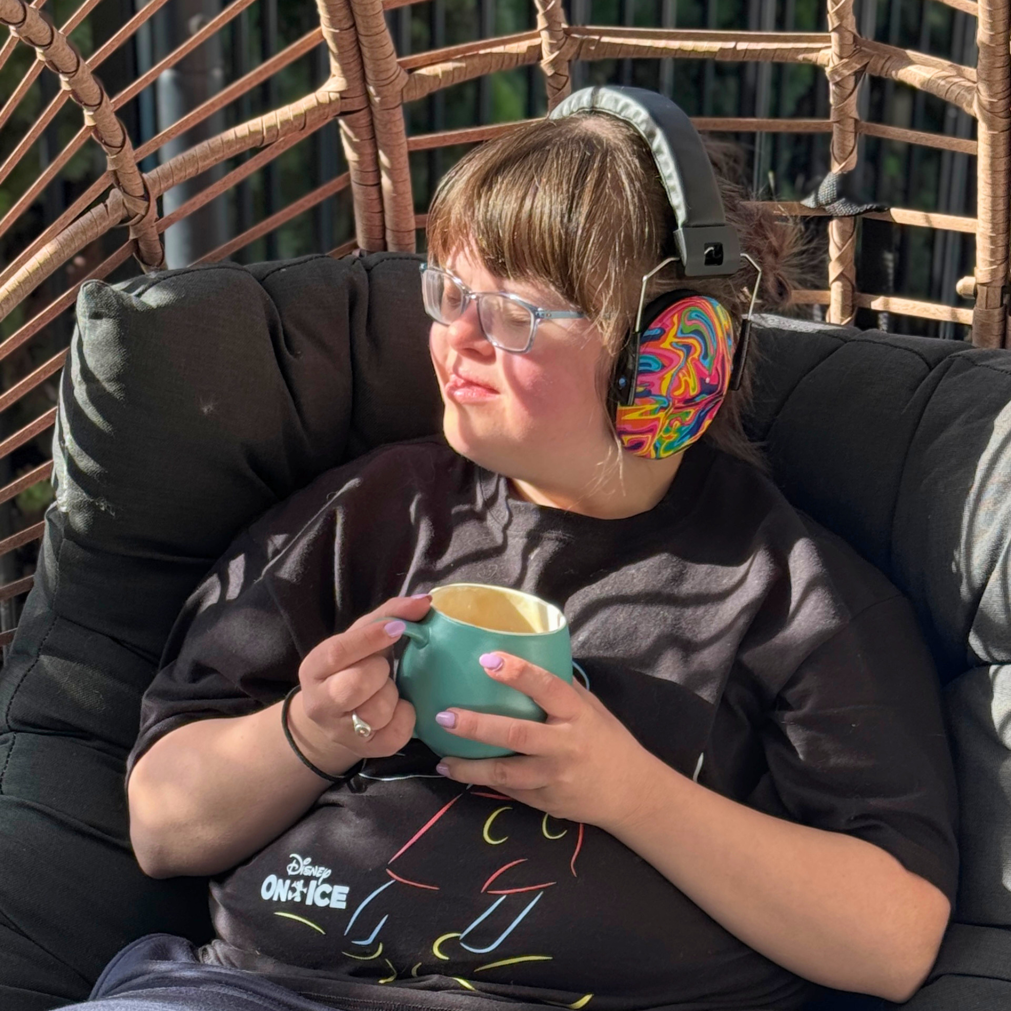 A person sitting in a wicker hanging chair outdoors, wearing colorful Starfish Noise Reduction Earmuffs and holding a teal mug. The person is dressed in a black graphic t-shirt and blue jeans, with a black cushioned chair behind them.