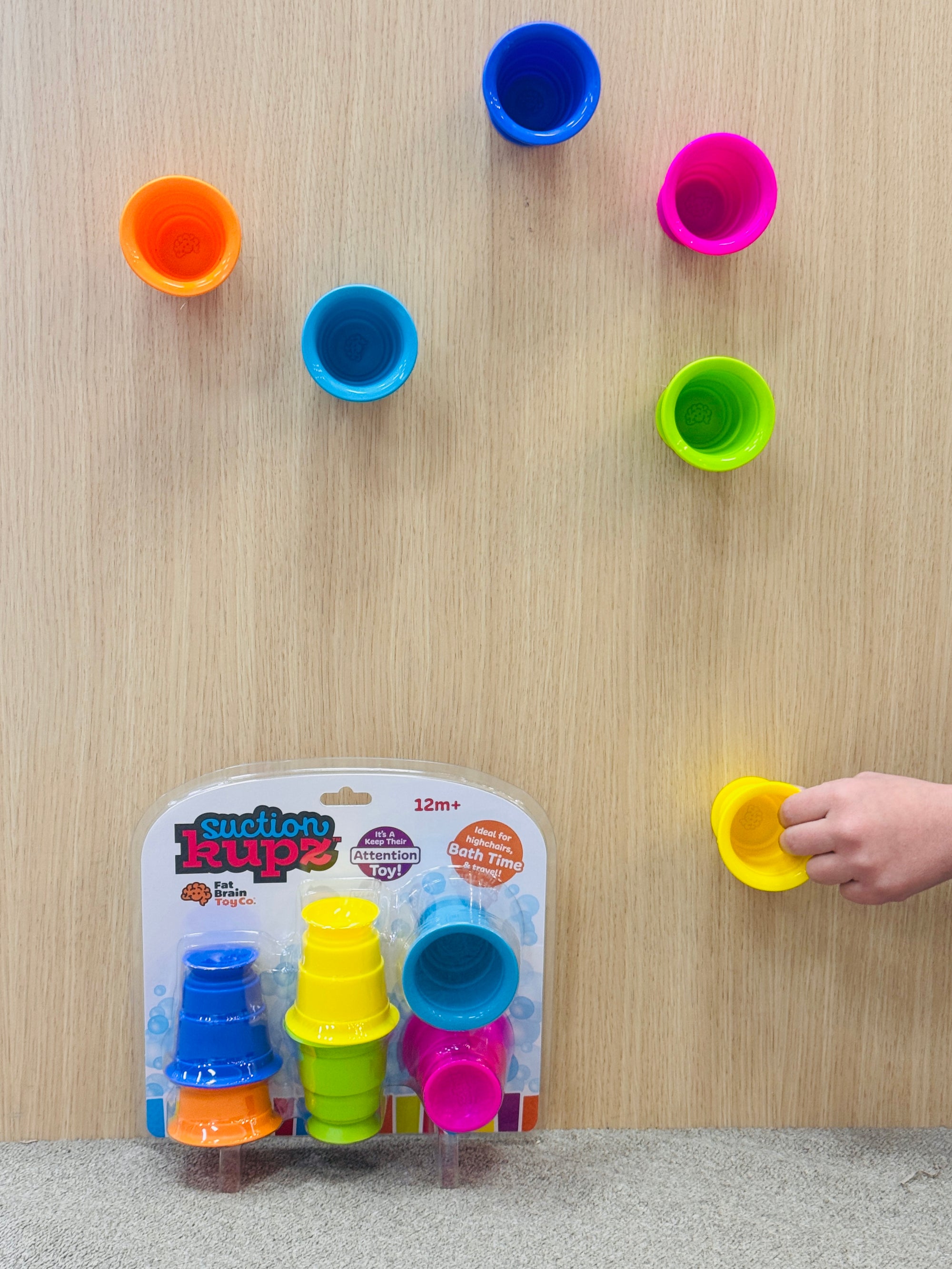 Fat Brain Suction Kupz on a wooden surface with a hand reaching for one, and the product packaging in the foreground.