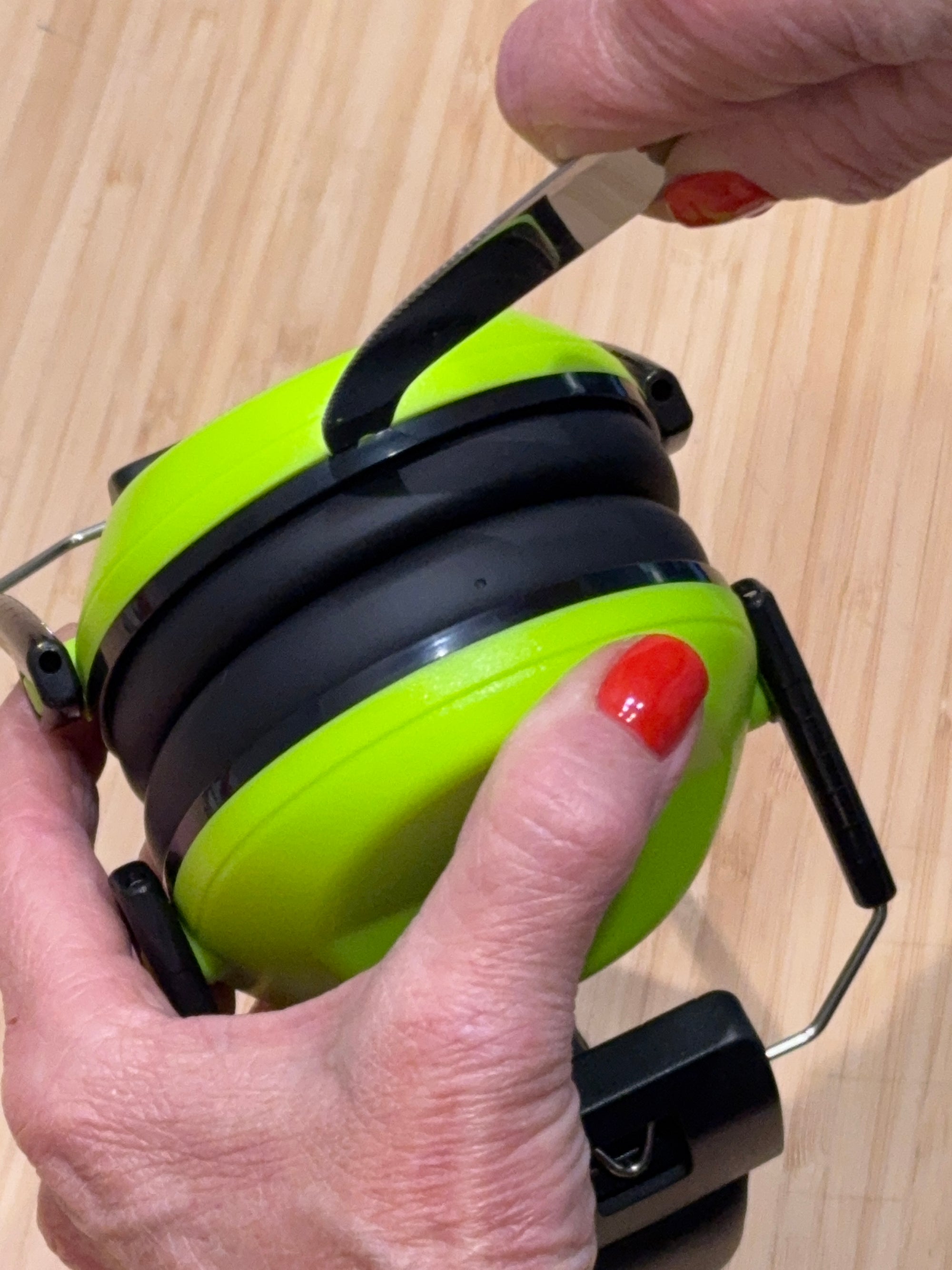 A close-up of a person holding a lime green and black over-ear hearing protection device, with a hand using a small tool to adjust or remove the black ear cup.