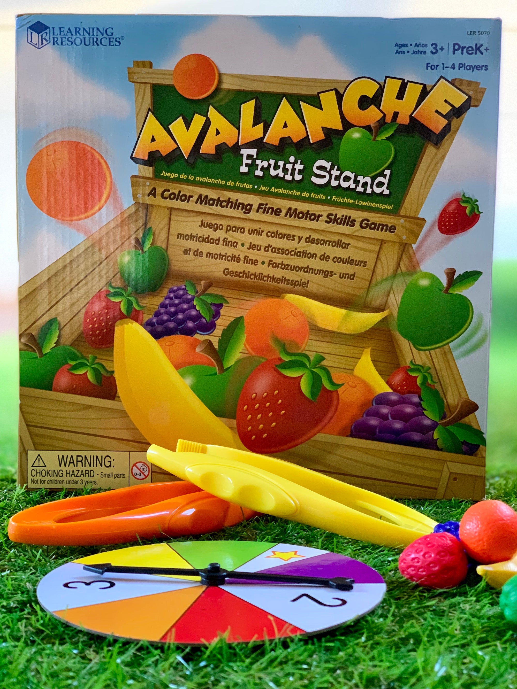 Avalanche Fruit Stand Game packaging box along with spinner and fruit laid out on grass